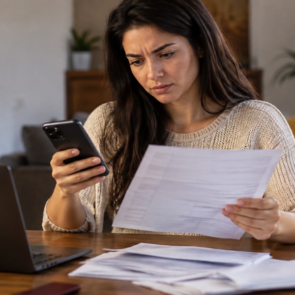 Pessoa em casa consultando o celular enquanto segura uma fatura, em um ambiente doméstico organizado e com expressão de atenção.